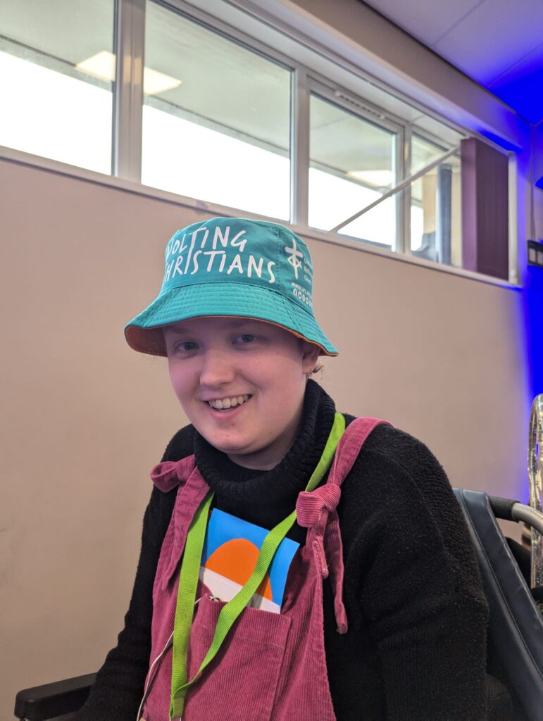photo of Sorrel in their electric wheelchair from above the waist. Sorrel is a young white person wearing pink cord dungarees over a black turtleneck, a green bucket hat with most of the words “Revolting Christians” and the United Reformed Church logo visible, a green lanyard, and their dungarees pocket is full of coloured voting cards. They’re smiling in a keen but tired way, as one would on day three of the United Reformed Church’s General Assembly.